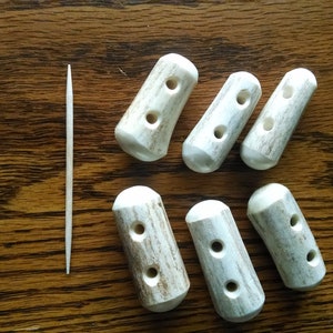 May include: Seven white antler bone toggles with two holes each. The toggles are arranged in a semi-circle on a wooden surface.