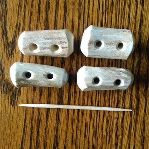 May include: Four light-colored, textured toggle buttons with two holes each, arranged in two rows. The buttons have a natural, mottled appearance with shades of beige and white. A thin, pointed wooden toothpick is positioned horizontally below them.