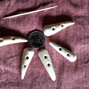 May include: Six bone buttons with two holes each. The buttons are a light cream color and are arranged in a star shape on a red fabric background.