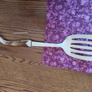 May include: A stainless steel spatula with a slotted head and an antler handle. The handle is a light brown color with a white tip. The spatula is lying on a purple floral patterned fabric.