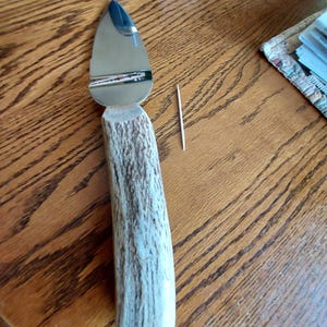 May include: A cheese knife with a polished steel blade and a handle made from a light-colored antler. A wooden toothpick is next to the knife. The knife is on a wooden surface.