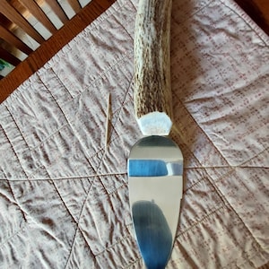 May include: A cake server with a handle made from a section of deer antler. The stainless steel blade has a blue gradient design. The server is resting on a quilted, patterned surface.