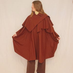 May include: A rust-colored cape coat with a layered design. The coat has a wide collar and a flowing silhouette. The person is wearing brown pants and brown heeled boots. The background is a plain white.