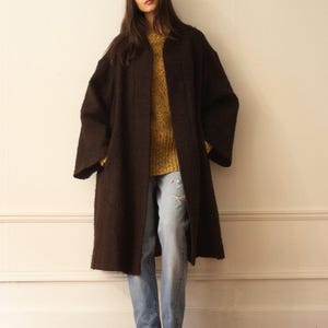 May include: A brown long-sleeved coat with a relaxed fit and a slightly oversized silhouette. The coat is worn over a yellow sweater and blue jeans.