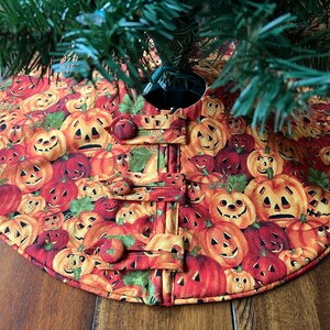 May include: A round, orange and red fabric tree skirt with a Halloween theme. The skirt is covered in a pattern of jack-o'-lantern faces. The skirt has a hole in the center for a tree trunk and is decorated with four buttons.