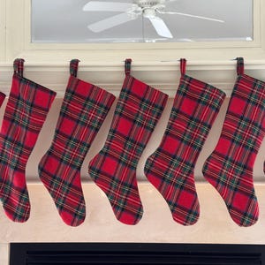 May include: A row of seven Christmas stockings with a red and green plaid pattern. The stockings are hanging from a white mantelpiece. The stockings are made of fabric and have a traditional Christmas design.