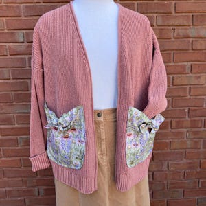 May include: A dusty rose cardigan with a long, open front and two large pockets. The pockets are lined with a floral fabric featuring a light purple, green, and white pattern.