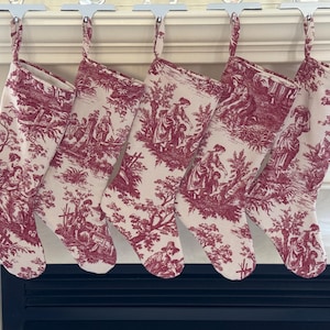 May include: Five Christmas stockings with a red and white toile pattern, hanging from silver hooks. The fabric features scenes of people in a landscape. The stockings are hanging above a black fireplace.