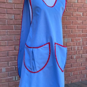 May include: A light blue apron with red trim and two large pockets. The apron has a wide neck opening and a long, flowing back.