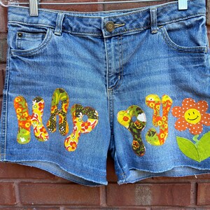 May include: Blue denim shorts with a patchwork design featuring the word "Happy" and a flower with a smiley face.