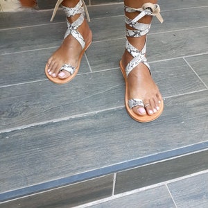 May include: A pair of brown leather sandals with a snake print design. The sandals have multiple straps that wrap around the ankle and tie at the top.