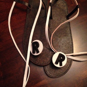 May include: Two brown leather sandals with white straps and black leather accents. Each sandal has a white circle with a black letter "R" on it.