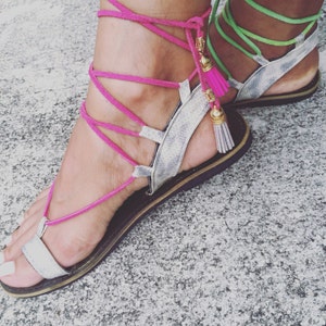Peut inclure: Sandales en cuir blanc avec des lacets et des glands roses et verts. Les sandales ont une semelle plate et sont attachées à la jambe.