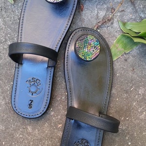 May include: A pair of black leather sandals with a decorative iridescent circle on the toe strap. The sandals have a simple strap design and a  "Lasco" logo on the sole.