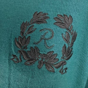 May include: A teal knitted jumper with a black embroidered wreath and a letter "R" in the centre.
