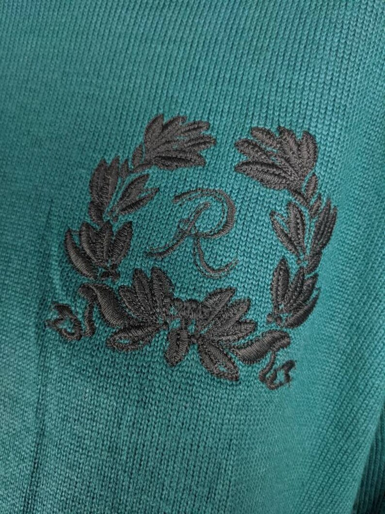 May include: A teal knitted jumper with a black embroidered wreath and a letter "R" in the centre.