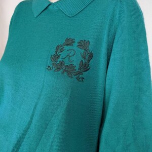 May include: Teal blue knitted sweater with a black embroidered logo of a letter "R" inside a wreath of leaves.