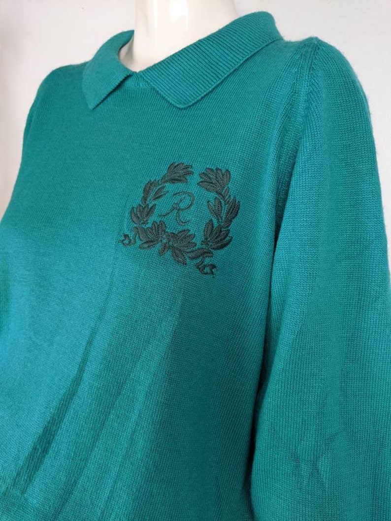 May include: Teal blue knitted sweater with a black embroidered logo of a letter "R" inside a wreath of leaves.