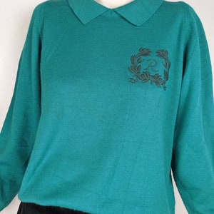 May include: Teal blue sweater with a collared neckline and a black embroidered logo on the front. The logo is a wreath with the letter "R" inside.