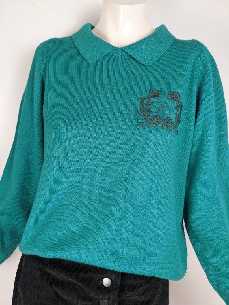 May include: Teal blue sweater with a collared neckline and a black embroidered logo on the front. The logo is a wreath with the letter "R" inside.