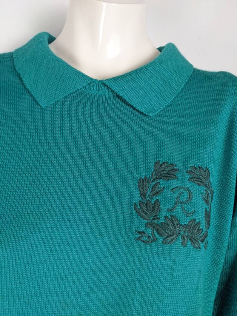 May include: Teal blue knitted sweater with a black embroidered wreath and the letter "R" in the centre.
