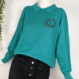 May include: A teal green sweater with a collared neckline and a black embroidered design on the front. The sweater has long sleeves and is made of a soft, knitted fabric.