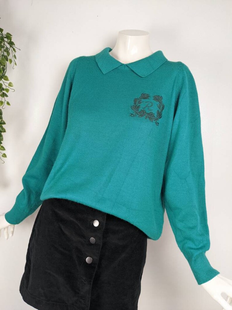 May include: A teal green sweater with a collared neckline and a black embroidered design on the front. The sweater has long sleeves and is made of a soft, knitted fabric.