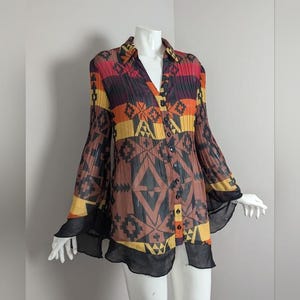 May include: A long-sleeved button-up shirt with a geometric pattern in shades of red, orange, yellow, and black. The shirt has a V-neck and bell sleeves with a black ruffle trim.