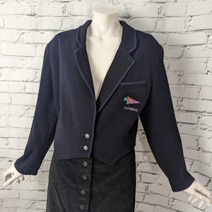 May include: A navy blue cropped blazer with a contrasting dark blue trim and a small embroidered patch that says "OUTRAGE". The blazer is worn over a black corduroy skirt with button closures.