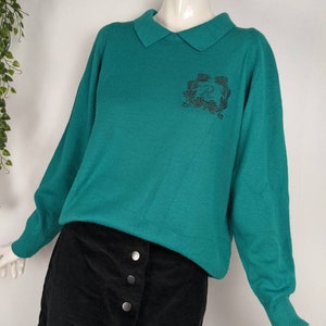 May include: Teal green sweater with a collared neckline and a black embroidered logo on the front. The sweater is worn with a black corduroy skirt.
