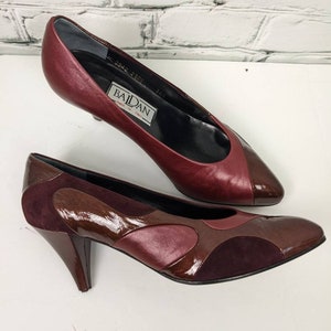 May include: Two pairs of women's dress shoes. The top shoe is a burgundy leather pump with a pointed toe. The bottom shoe is a burgundy, brown, and dark purple leather pump with a unique design and a curved heel. The shoes are on a white surface.