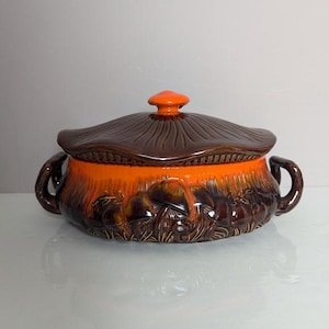 May include: A brown and orange ceramic casserole dish with a lid shaped like a mushroom. The dish is decorated with a mushroom motif.