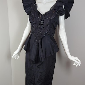 May include: A black vintage dress with a peplum waist and shoulder ruffles. The bodice has a V-neck with sequin and bead detailing. The dress, displayed on a mannequin, has a knee-length skirt.
