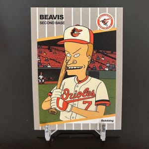May include: A baseball card featuring a cartoon character wearing a Baltimore Orioles uniform. The card has the text "BEAVIS SECOND BASE" and "BALTIMORE ORIOLES" on it.