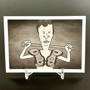 May include: Black and white cartoon print of a character holding a baseball bat, wearing shoulder pads. The image is displayed in a white frame and propped up on a clear stand.