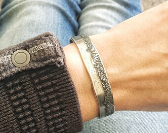 Könnte beinhalten: Ein silbernes Armband mit einer strukturierten, gehämmerten Oberfläche. Das Armband wird am Handgelenk getragen.