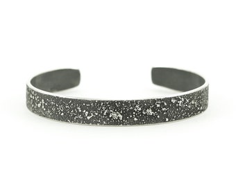 Könnte beinhalten: Ein silbernes Armband mit einer strukturierten, gesprenkelten Oberfläche. Das Armband ist ein einfaches, offenes Band.