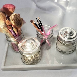 May include: A white marble tray with four glass jars containing makeup brushes, cotton swabs, and a pink eyebrow razor. The jars have silver lids.