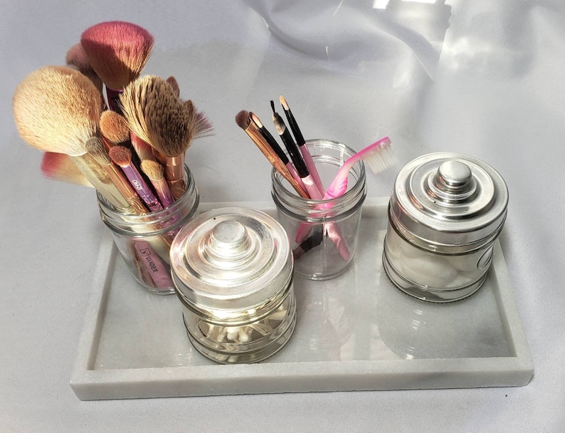 May include: A white marble tray with four glass jars containing makeup brushes, cotton swabs, and a pink eyebrow razor. The jars have silver lids.