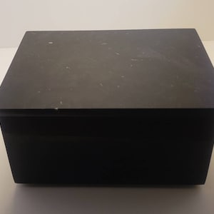 May include: A black rectangular box with a lid. The box is made of a smooth, matte material.