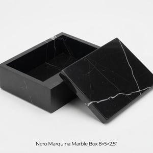 May include: A black Nero Marquina marble box with a matching lid. The box is rectangular, with the lid slightly ajar. White veining patterns the polished black surface. The box measures 8x5x2.5 inches.