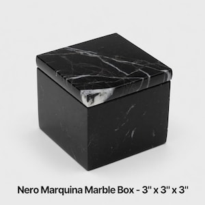 May include: A square Nero Marquina marble box with a removable lid, featuring a black base colour and white veining throughout. The box measures 7.62 cm by 7.62 cm by 7.62 cm as indicated by the text printed below the object in the image.