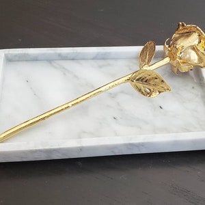 May include: A gold-plated rose on a white marble tray. The rose is a single stem with a single bloom and leaves.