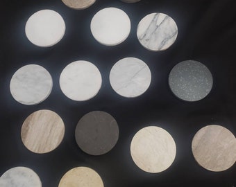 Può includere: Una collezione di 14 sottobicchieri rotondi in vari colori e motivi, tra cui bianco, grigio, marrone e nero. I sottobicchieri sono disposti in un motivo circolare su una superficie nera.