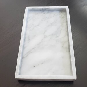 May include: A white marble tray with a raised border. The tray is rectangular and has a smooth surface.