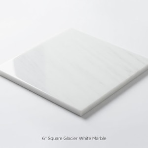 May include: A square, 15 cm Glacier White marble tile. The tile is a bright white with subtle grey veining, and has a smooth, polished surface. The text "6" Square Glacier White Marble" is at the bottom of the image.