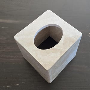 May include: A square tissue box with a light beige marble-like finish. The box has a large oval opening on the top. The tissue box is a decorative item for the home.