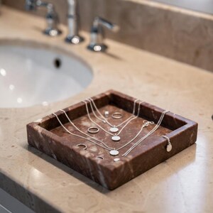 May include: A square, reddish-brown marble tray containing several silver-toned necklaces with circular pendants and multiple silver rings. The jewelry is arranged neatly within the tray, which features natural stone veining and a polished finish.