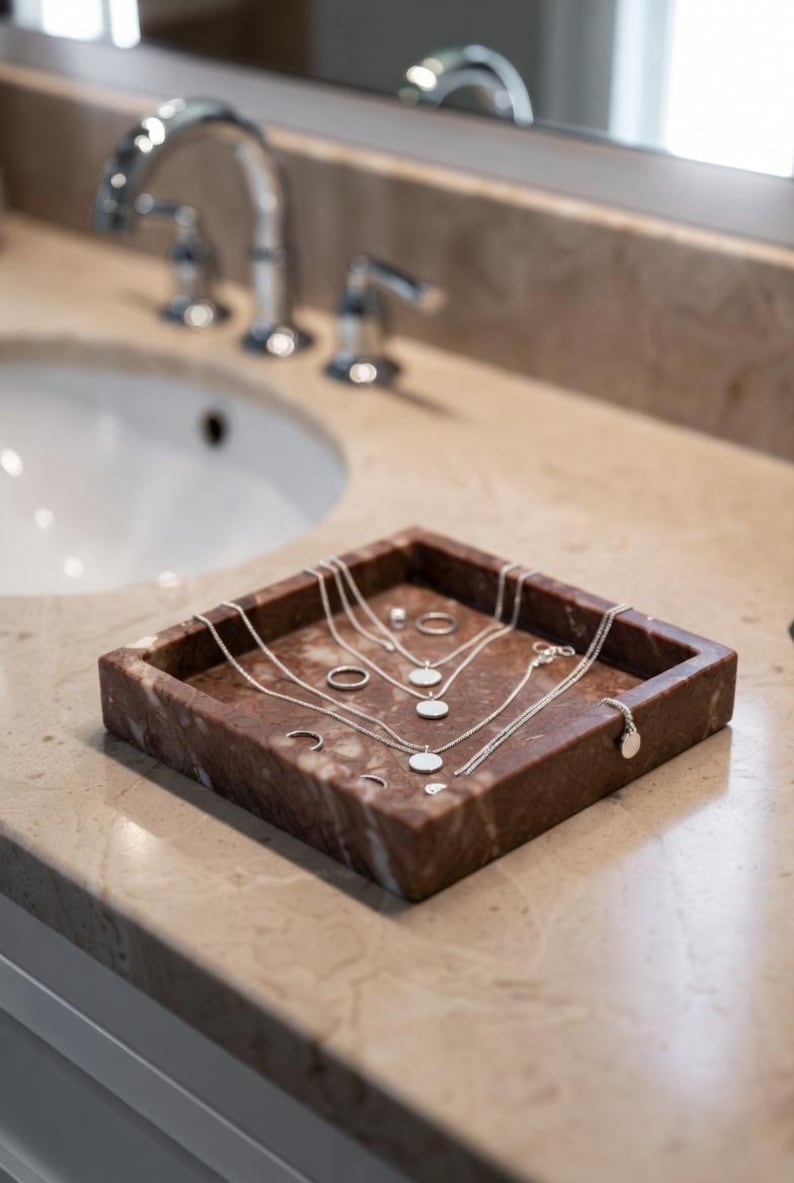 May include: A square, reddish-brown marble tray containing several silver-toned necklaces with circular pendants and multiple silver rings. The jewelry is arranged neatly within the tray, which features natural stone veining and a polished finish.