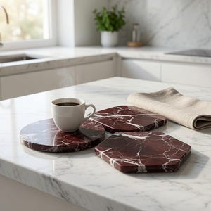 May include: A white ceramic coffee cup filled with dark coffee sits on a round, deep red marble coaster with white veining. Nearby are a square and a hexagonal coaster, both made of the same red marble with white veining, and a folded beige cloth.